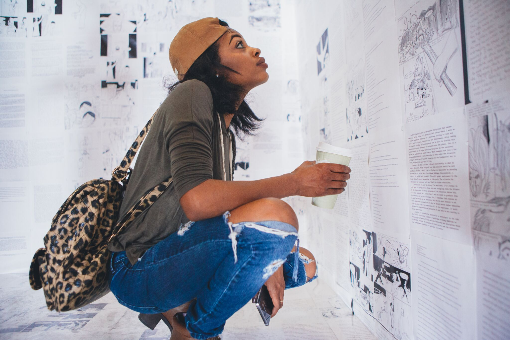 From her right side, a woman wearing jeans, an olive green long-sleeved shirt, sleeves pushed up past her elbow, a leopard-print backpack, and a tan cap, squats and looks intently upwards and towards the right, towards the artwork - black and white text, images, and drawings - like pages of a book - plastered against the wall in front of her behind her, and reflected on the white floor below her.