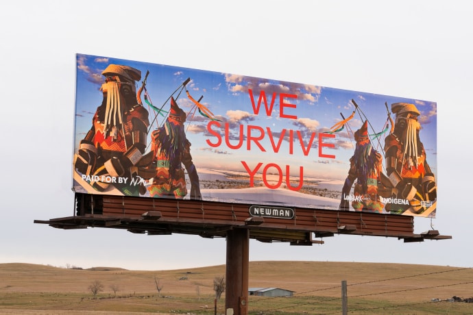 A colorful billboard stands in brownish ranchland. On the billboard, backed by an image of somewhat cloudless blue sky, two figures in colorful clothing reminiscent of armored beadwork are vertically mirrored to create four figures flanking three words at the center of the billboard in all caps and red 