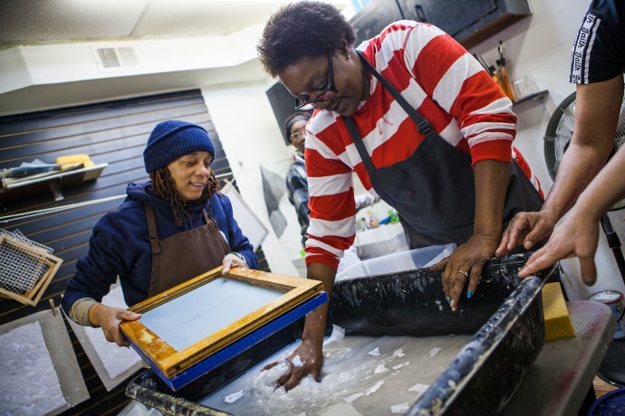 In a busy print shop, a figure in a blue apron and red-and-white horizontally striped shirt and glasses mixes cloudy white liquid with paper fragments in a large black tub with their right hand as another wearing a blue sweatshirt, blue knit cap, and brown apron holds a screenprinting rig above the surface of the liquid, readying to place it in the tub.