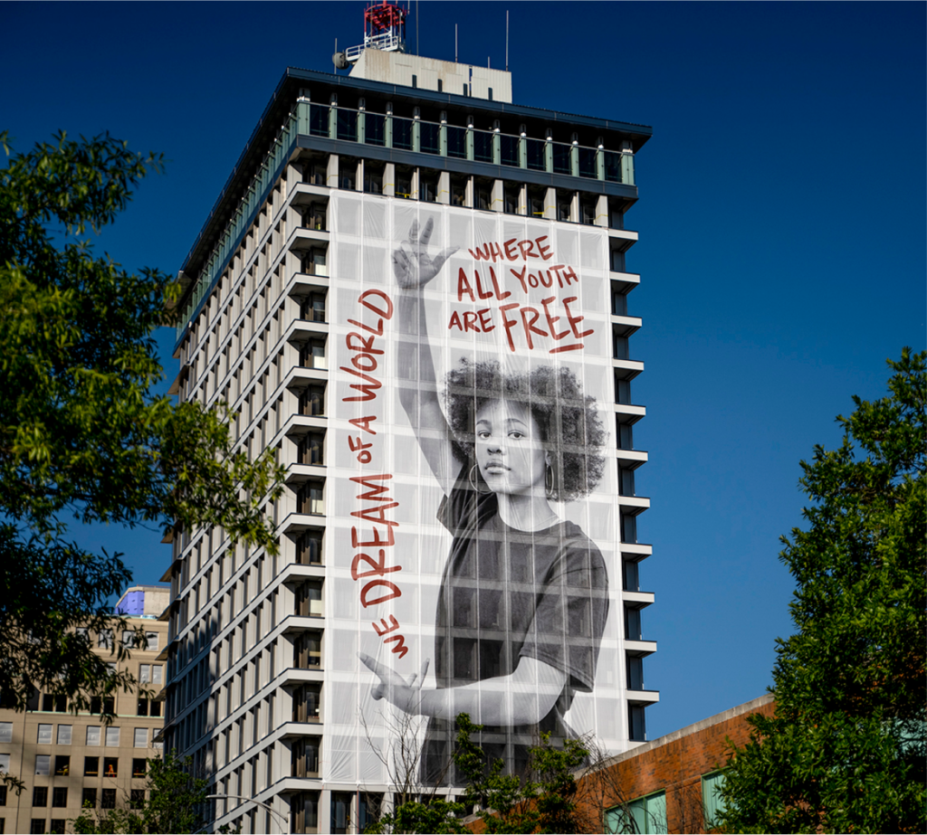 On the side of a building at least 15 stories high is a 12-story translucent white banner with the black-and-white image of a young woman in a t-shirt turned to look at you expectantly with her right arm raised, thumb and first two fingers extended, and left arm across her waist, hand open upward. Extending from her open left hand up and over her are the words, in red, 