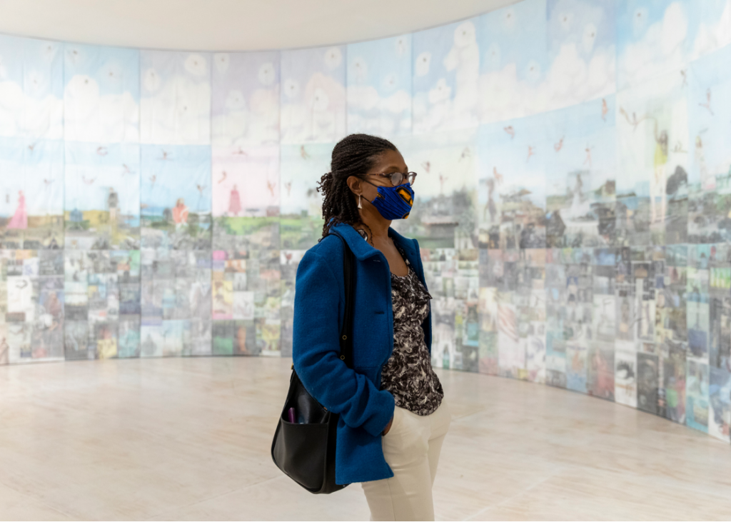 A4J Advisor Tanya Coke stands facing right in a blue jacket, blue face mask, white pants, black-and-white blouse and black shoulder bag, in front of the large art piece, extending in an arc behind and presumably to her right. The work is a series of pastel colored column-like panels, 11 in view, with cloud-like feel near the top towards the ceiling edge, leading downward into more detailed squarish, darker pastel image blocks at the floor edge.