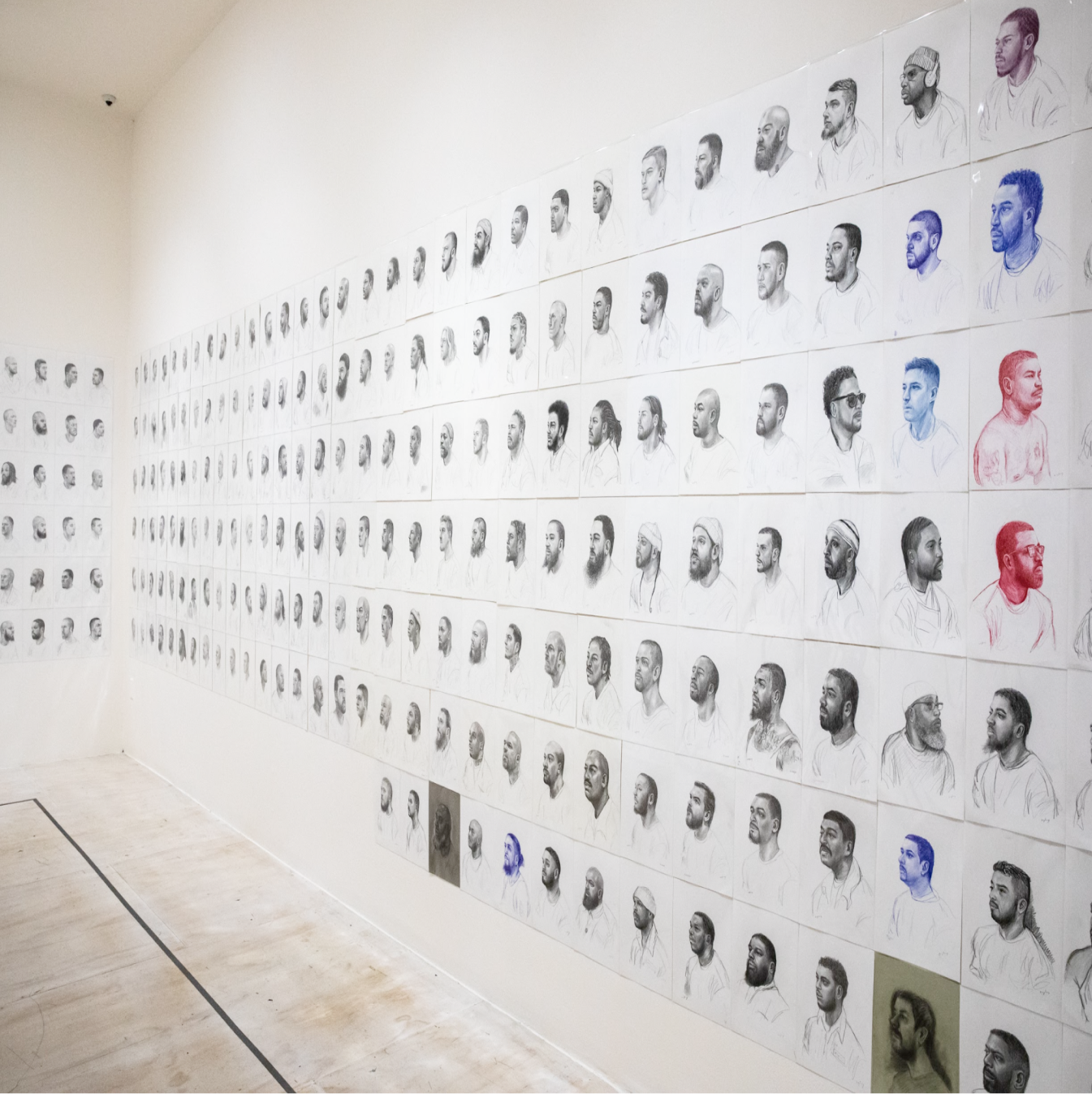 A white wall extends from right to left, turning a corner, covered with seven and then six rows of individual portraits. From neck above, facial features are shaded in detail largely in black and grays (with a few in red or blue). Over 200 individual portraits of are in view.