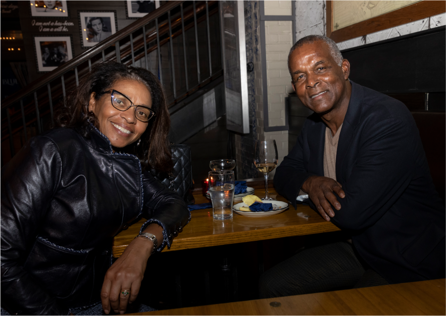 In a restaurant, two figures sit across from one another at a small table with drinks and appetizers. The woman on the left wears glasses, a black jacket, leans her left elbow on the table and turns with a smile at you. The man on the right wears a tan shirt, dark blue jacket and leans slightly forward with a smile at you, arms folded on the table.
