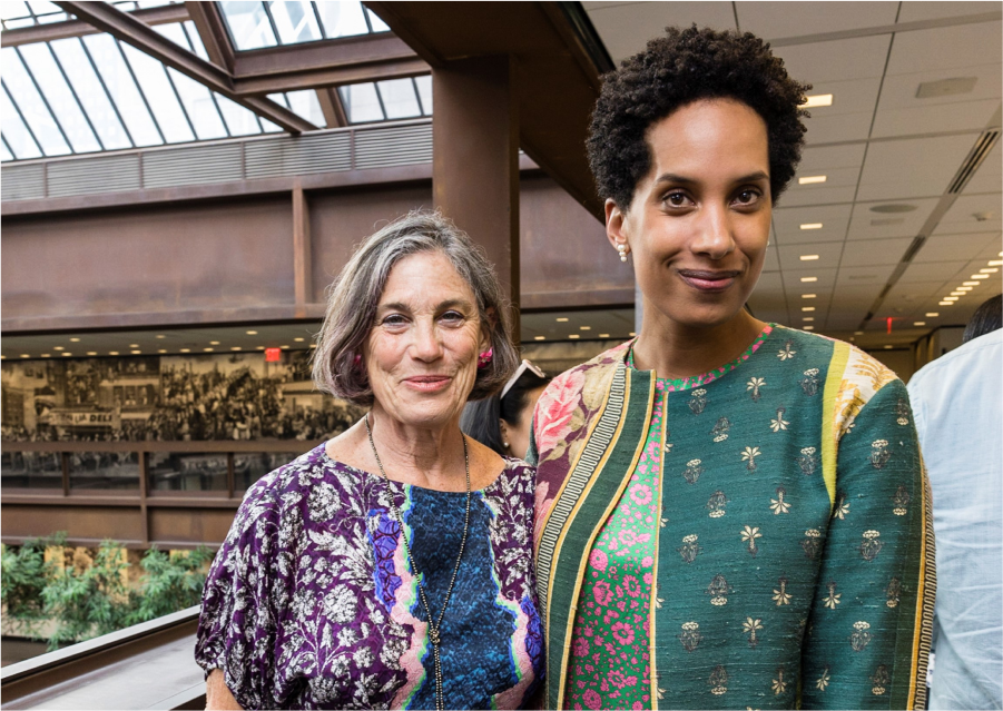 Two women stand, look, and smile towards you, arm and arm with each other in the interior balcony walkway of an office building. On the left, she wears a patterned purple floral dress. On the right, she wears an overcoat over a bright pink and green floral dress.